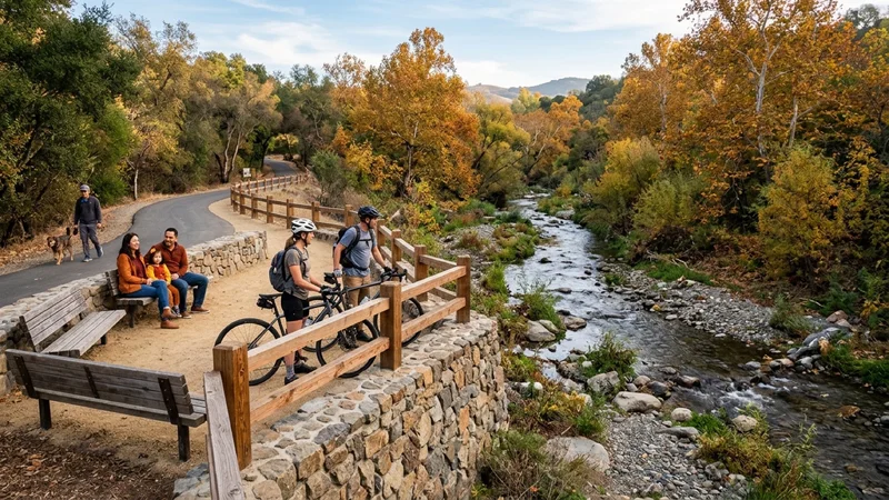 Cyclists and walkers gathered at a stone-and-timber creekside overlook deck beside a restored cobble stream channel and autumn sycamores along the Arroyo Mocho Trail in Pleasanton