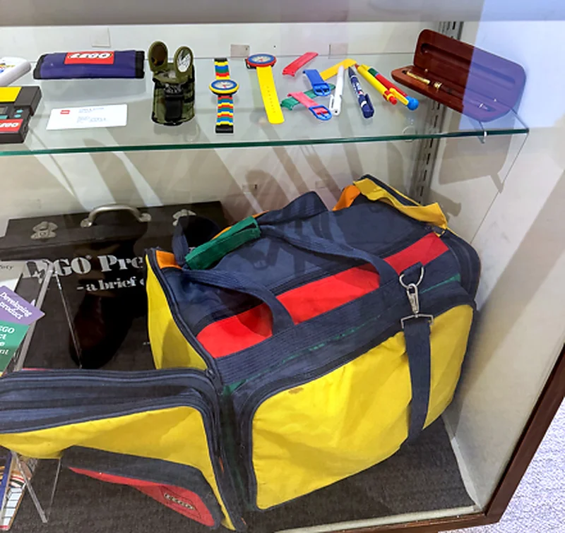 A primary-colored vintage LEGO travel duffel bag displayed beneath a shelf of archival LEGO watches, pens and a wooden pen case.