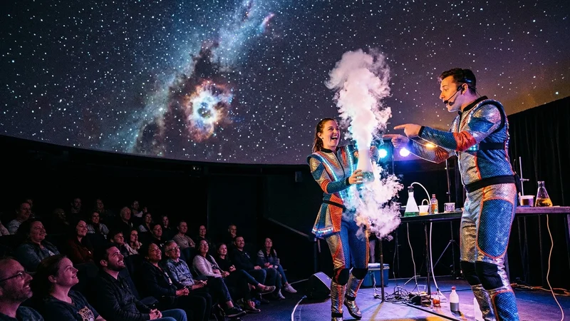 Performers in futuristic costumes conducting a dry-ice experiment on stage beneath a planetarium dome in Oakland
