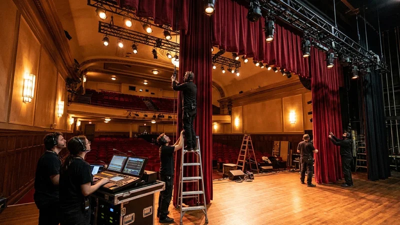 Theater technical directors and stagehands adjusting crimson velvet acoustic stage curtains and modern LED spotlights inside the historic Village Theatre in Danville